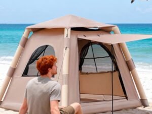 A man sitting in front of a beach tent with an open flap on a sandy beach near the ocean.